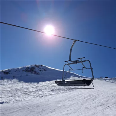 A ski lift hovers over a snow-covered slope under a clear blue sky. The sun shines brightly in the sky.