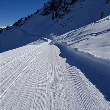 A snow-covered ski slope with fresh tracks in the snow. In the background, mountainous landscapes and a blue sky can be seen.