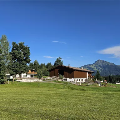 A picturesque landscape with a large house and green meadows. In the background, mountains stretch under a clear blue sky.