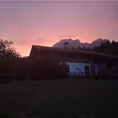 A house at twilight with a colorful sky. In the background, majestic mountains and trees can be seen.