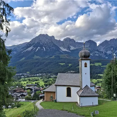 A picturesque church stands in a quiet village, surrounded by green meadows. In the background, majestic mountains rise under a partly cloudy sky.