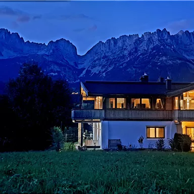 A modern house at dusk, surrounded by majestic mountains. The soft lights of the house shimmer in the serene landscape.
