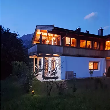 A modern house with cozy lights that shines in the twilight. In the background, gentle hills and a clear evening sky can be seen.