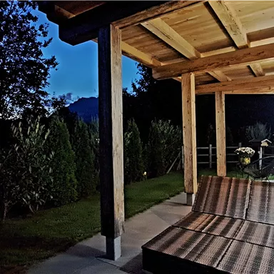 A cozy porch with a wooden ceiling under a clear night sky. Nearby, plants and trees are visible.