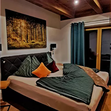 A cozy bedroom with a large bed and modern pillows. The wooden beam ceiling and the artistic picture on the wall give the room a warm atmosphere.