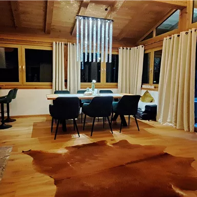 A modern living room with wooden floors and large windows. The space is elegantly furnished and features a dining area with a table and chairs.