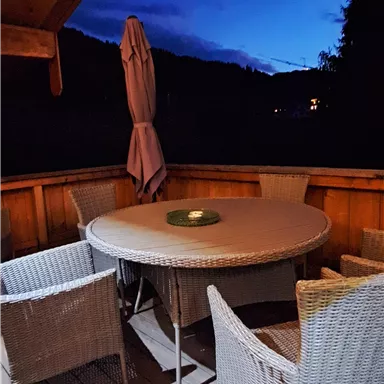 A cozy outdoor area with a round table and chairs made of rattan. The sky is dark blue, indicating the evening.