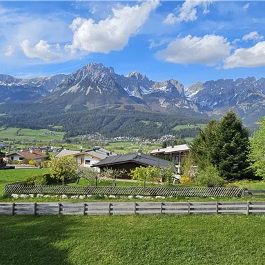 A picturesque landscape with green meadows and an impressive mountain range in the background. The sky is clear and blue with some clouds.