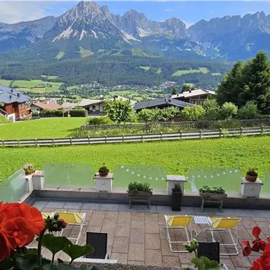 A picturesque mountain landscape with green meadows and rugged peaks. In the foreground, blooming geraniums on a terrace.
