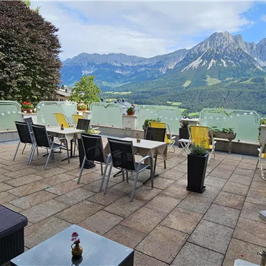 A cozy terrace with tables and chairs offers a breathtaking view of the mountains. The surroundings are green and inviting, perfect for relaxing.