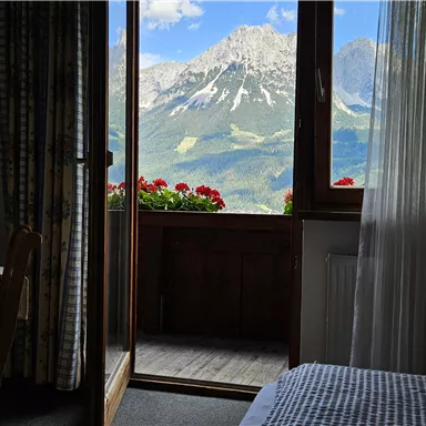 A cozy room with a view of the mountains and a balcony full of flowers. The view shows a picturesque landscape with a blue sky and clouds.