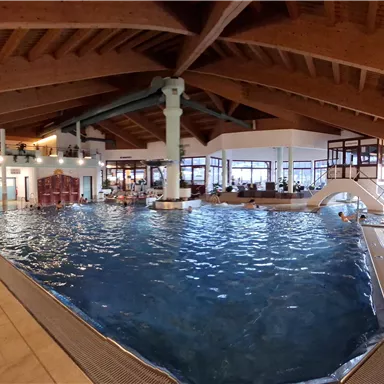 A large indoor swimming pool with clear water and a wooden ceiling. Surrounded by cozy loungers and seating areas for guests.