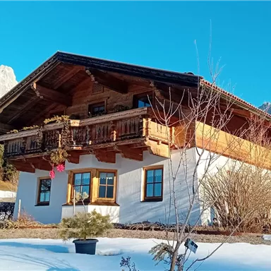 A traditional Alpine house in the snow with a beautiful balcony. In the background, majestic mountains and a clear blue sky are visible.