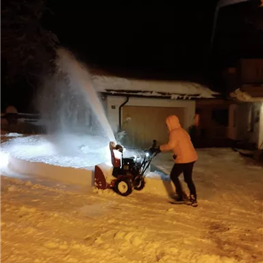 A person is operating a snow blower in a snowy environment at night. The snow is being blown into the air, and a brightly lit house can be seen in the background.