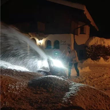 A snowplow is clearing snow at night in front of a house. The snow is being pushed away with bright light.