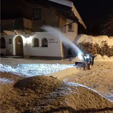 A man is clearing snow with a snow plow at night. In the background, a chalet-like house and a snowy garden can be seen.