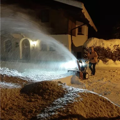 A person is clearing the snow-covered path in front of a house with a snow blower. It is nighttime, and the light from the machine illuminates the surroundings.