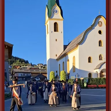 A procession in traditional costume passes by a church. The sun is shining and the sky is blue.