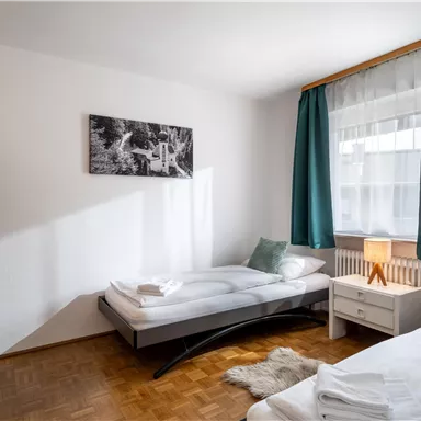 A simple bedroom with two single beds and a bright atmosphere. Large windows and green curtains create a cozy space.