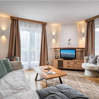 A modern living room with a cozy sofa, a wooden table, and a television. Large windows let in a lot of light and give the room an inviting atmosphere.