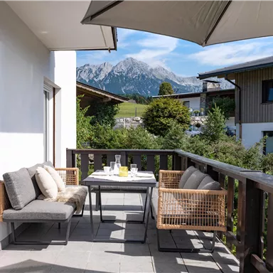 A cozy terrace with a table and chairs, surrounded by a beautiful mountain landscape. The sun is shining and the sky is blue.