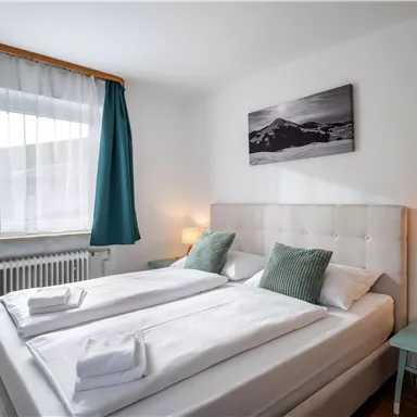 A cozy bedroom with a large bed and two pillows. The walls are light, and the window is decorated with curtains in blue and white.