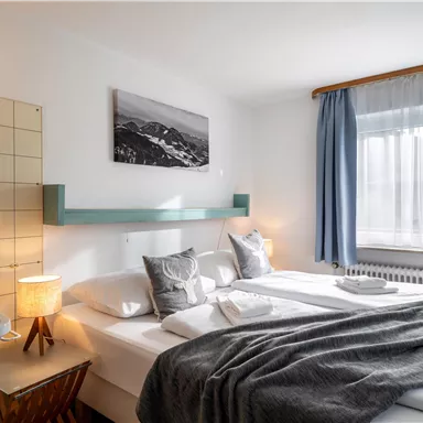 A cozy bedroom with a double bed and two pillows. The room has large windows and a calm, friendly atmosphere.