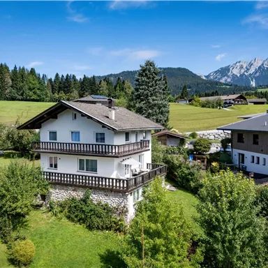 A beautiful house in a rural area, surrounded by green fields and trees. In the background, mountains and a blue sky can be seen.
