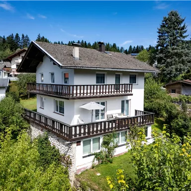 A cozy house with several balconies, surrounded by trees and meadows. In the background, mountains and a clear blue sky can be seen.
