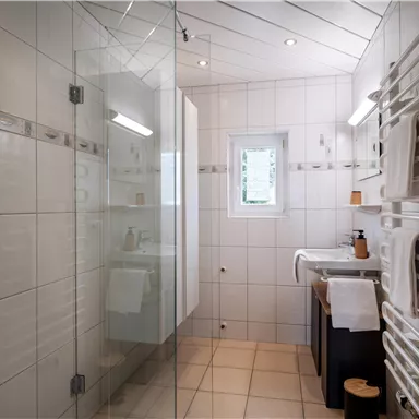 A modern bathroom with a glass shower and bright tiles. On the wall, there is a towel warmer, and a large window provides daylight.