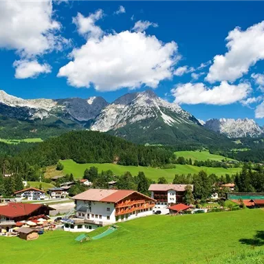 A picturesque mountain landscape with green meadows and majestic mountains in the background. In the foreground, traditional alpine houses can be seen under a radiant blue sky.