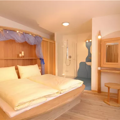 Ein modernes Schlafzimmer mit einem großen Doppelbett und Holzoberflächen. Im Hintergrund befindet sich ein angrenzendes Badezimmer mit Dusche.