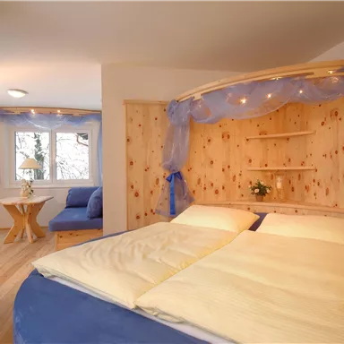 Ein gemütliches Schlafzimmer mit einem großen Bett und hellblauer Bettwäsche. Im Hintergrund sind Fenster und eine kleine Sitzecke sichtbar.