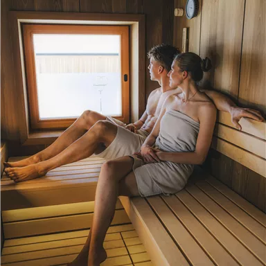 A couple sits relaxed on a wooden bench in a sauna. The large window front provides plenty of daylight.