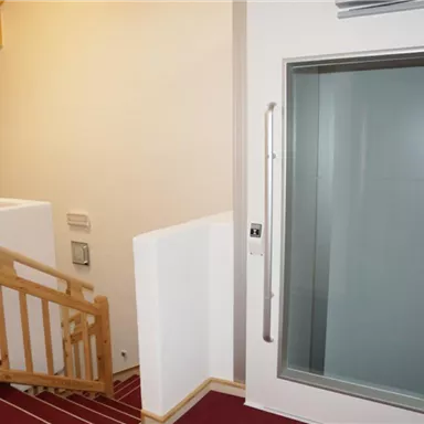 Ein moderner Innenraum mit einer Treppe und einem Glasaufzug. Der Boden ist mit rotem Teppich ausgelegt und die Wände sind hell.