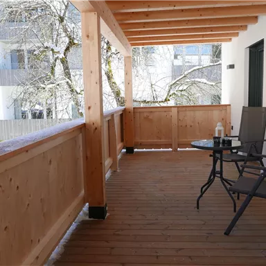 A cozy balcony with wood cladding, furnished with a table and chairs. In the background, trees and a bright residential complex can be seen.