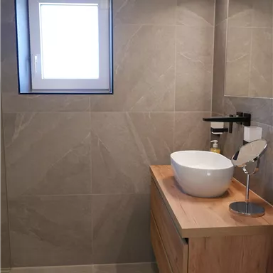 A modern bathroom with gray tiles and a wooden sink unit. The round sink and the large mirror create a bright, friendly atmosphere.