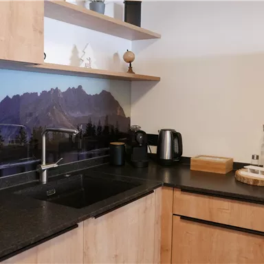 A modern kitchen with wooden shelves and a dark countertop. In the background, mountains and trees are depicted.