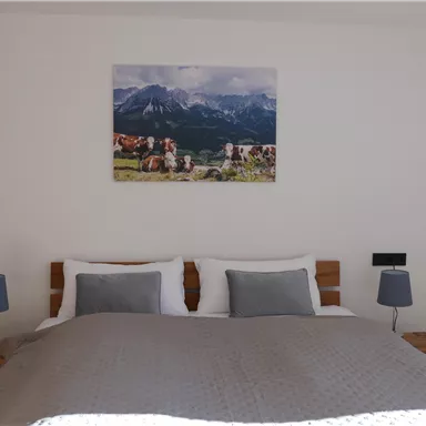 A cozy bedroom with a large bed, two nightstands, and lamps. On the wall hangs a picture of a mountain landscape with cows.