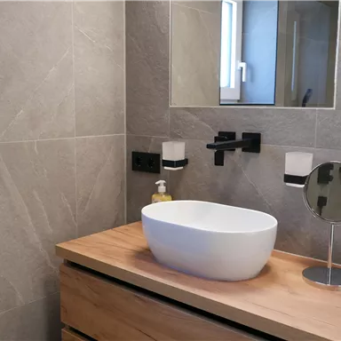 A modern bathroom with gray tiles and an elegant wooden vanity. In the foreground is a round sink and a mirror.