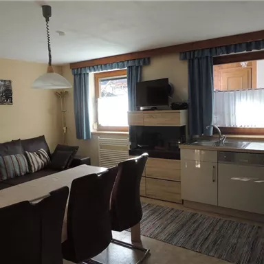 A cozy living kitchen with a dining table and a comfortable sofa area. The windows are equipped with blue curtains, and the room is bright and inviting.