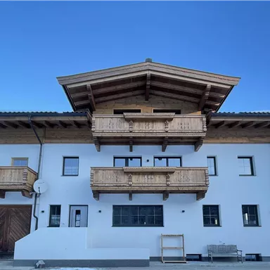 A modern, two-story house in alpine style with wooden balustrades. The sky is clear and blue, and the ground is covered with snow.