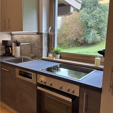 Eine moderne Küche mit einem großen Fenster, das auf einen grünen Garten blickt. Auf der Arbeitsplatte befinden sich Herd und Spüle.