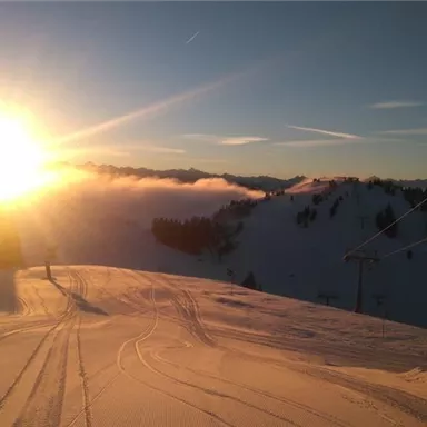 A snowy mountain landscape at sunset. The sky is clear and the sun shines over the hills.