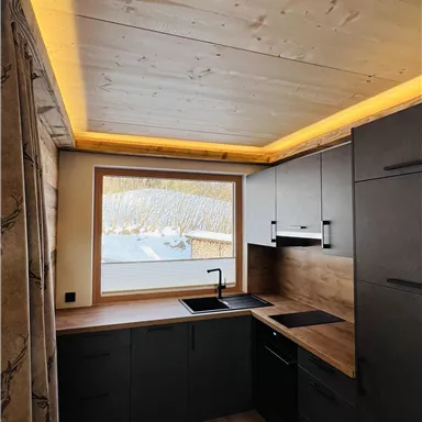A modern kitchen with a wooden decor and a large window. Soft lighting on the ceiling creates an inviting atmosphere.