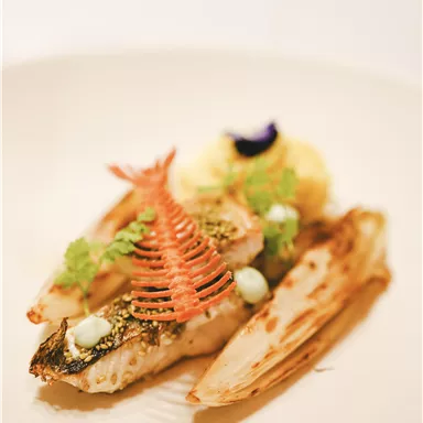 A artfully arranged fish on a white plate, garnished with fresh herbs and colorful decorations. Next to it, grilled vegetable sides can be seen.