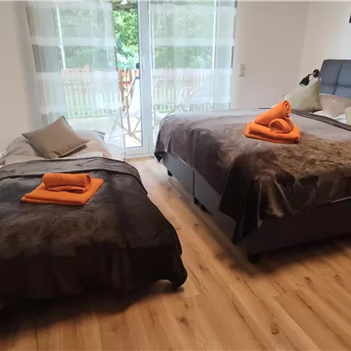 Ein gemütliches Schlafzimmer mit zwei Betten, einem großen Fenster und Holzfußboden. Orangefarbene Handtücher liegen ordentlich auf den Bettdecken.