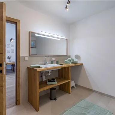 A modern bathroom with a wooden vanity and a large mirror. There are green towels and a bright, minimalist style.