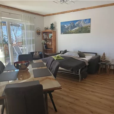 Ein helles und einladendes Wohnzimmer mit einem Esstisch und Stühlen. Im Hintergrund ist ein gemütliches Bett und ein Fenster mit Blick auf die Natur.