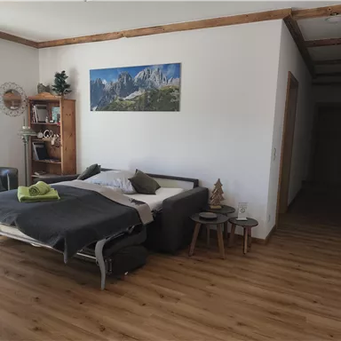 Ein gemütliches Zimmer mit einem Bett und einem Sofa. An der Wand hängt ein Landschaftsbild, und der Raum hat Holzfußboden und helle Wände.